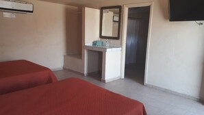 Quarto de hotel com duas camas de solteiro, um banheiro pequeno com espelho e uma TV fixada na parede.