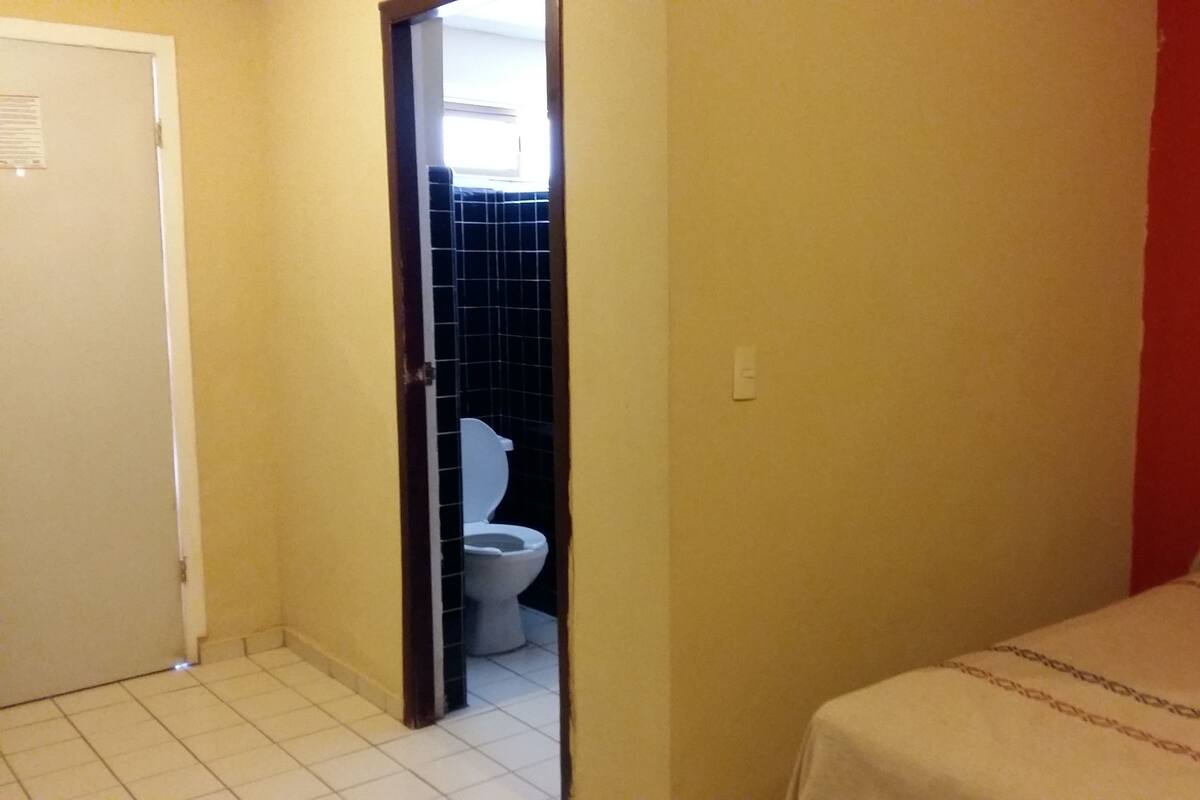 Une petite pièce étroite, avec un lit, une porte blanche et une vue sur une salle de bain équipée de toilettes et dont un mur est recouvert de carreaux.