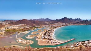 Uma vila costeira com uma praia, águas azul-claras e montanhas ao fundo.