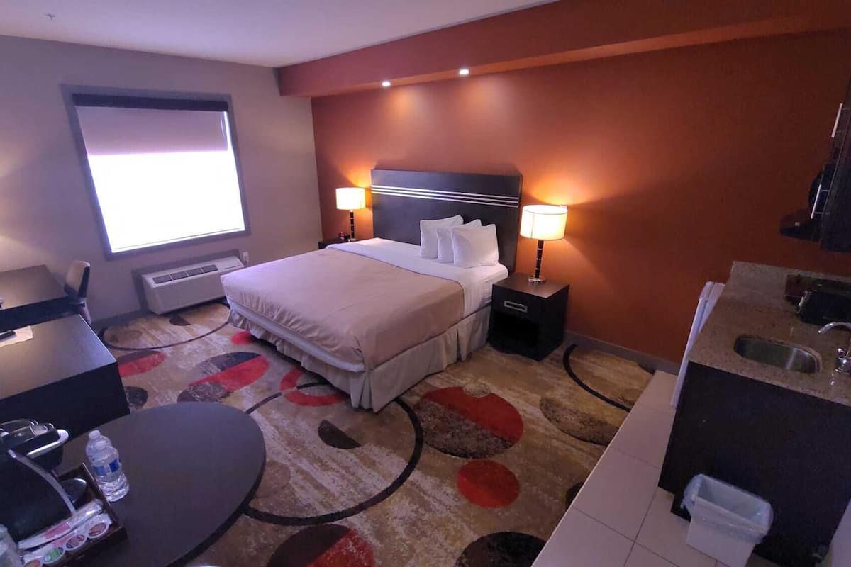 A hotel room with a bed, two bedside lamps, a desk with a chair, a small table with a chair, a window, and a bathroom with a sink and mirror.