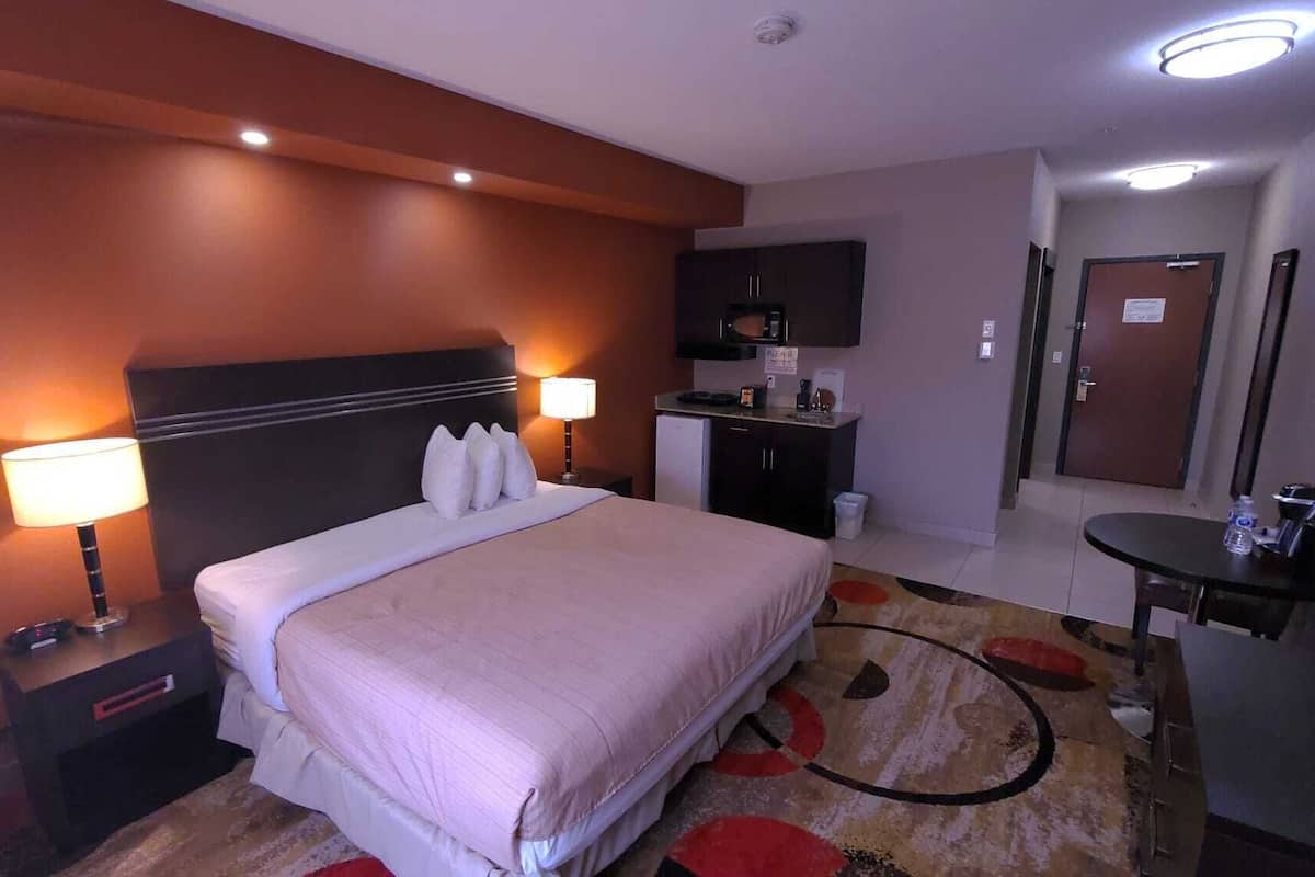 A hotel room with a bed, bedside lamps, a desk, a small kitchenette, and a door.