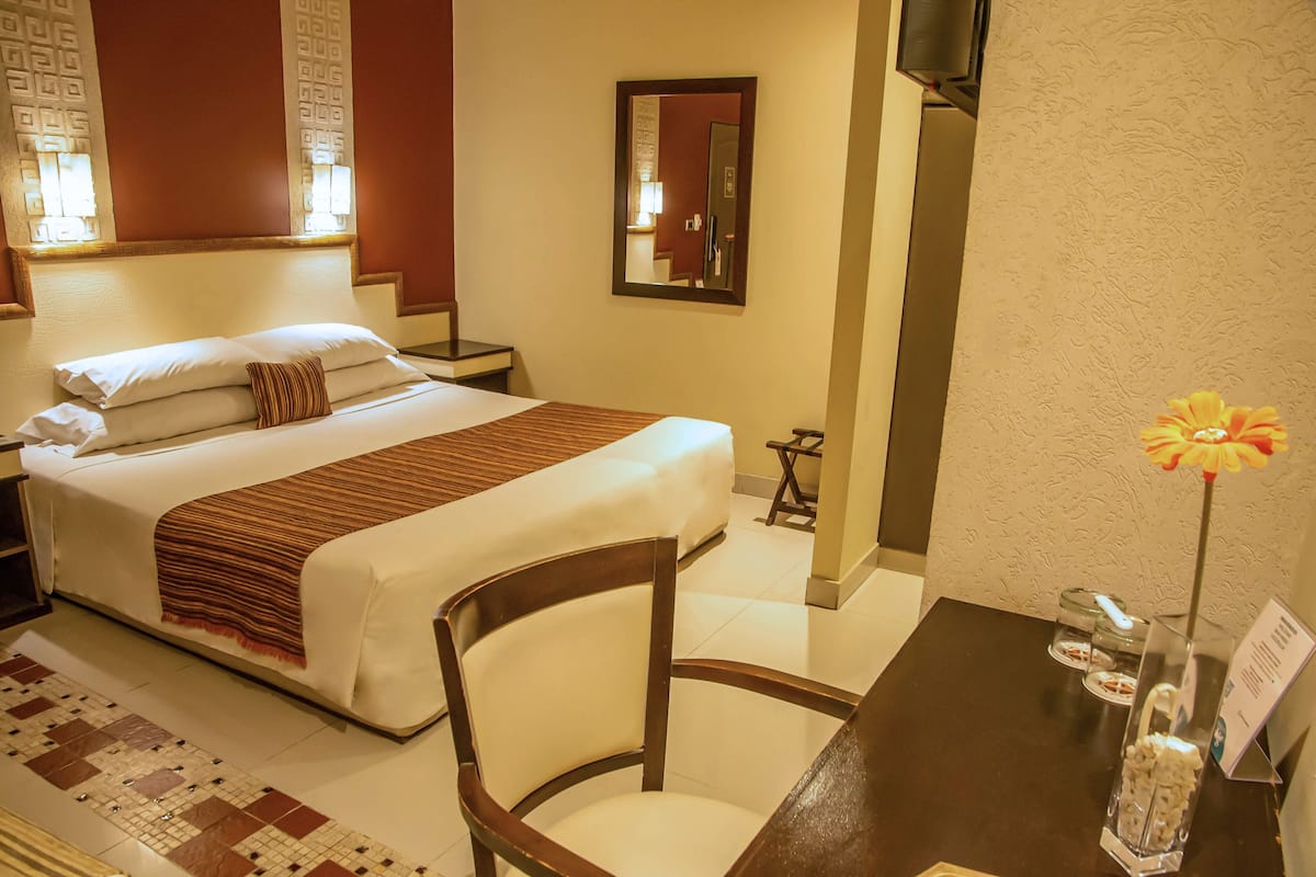 Habitación de hotel con una cama bien hecha, una silla, una mesita con un florero y un espejo en la pared.
