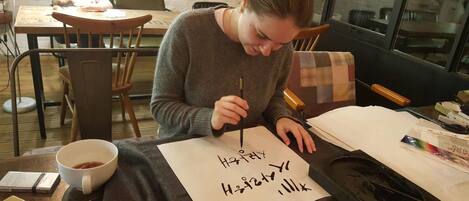 A person writing on a piece of paper at a table with a cup of tea, a smartphone, and some writing materials.
