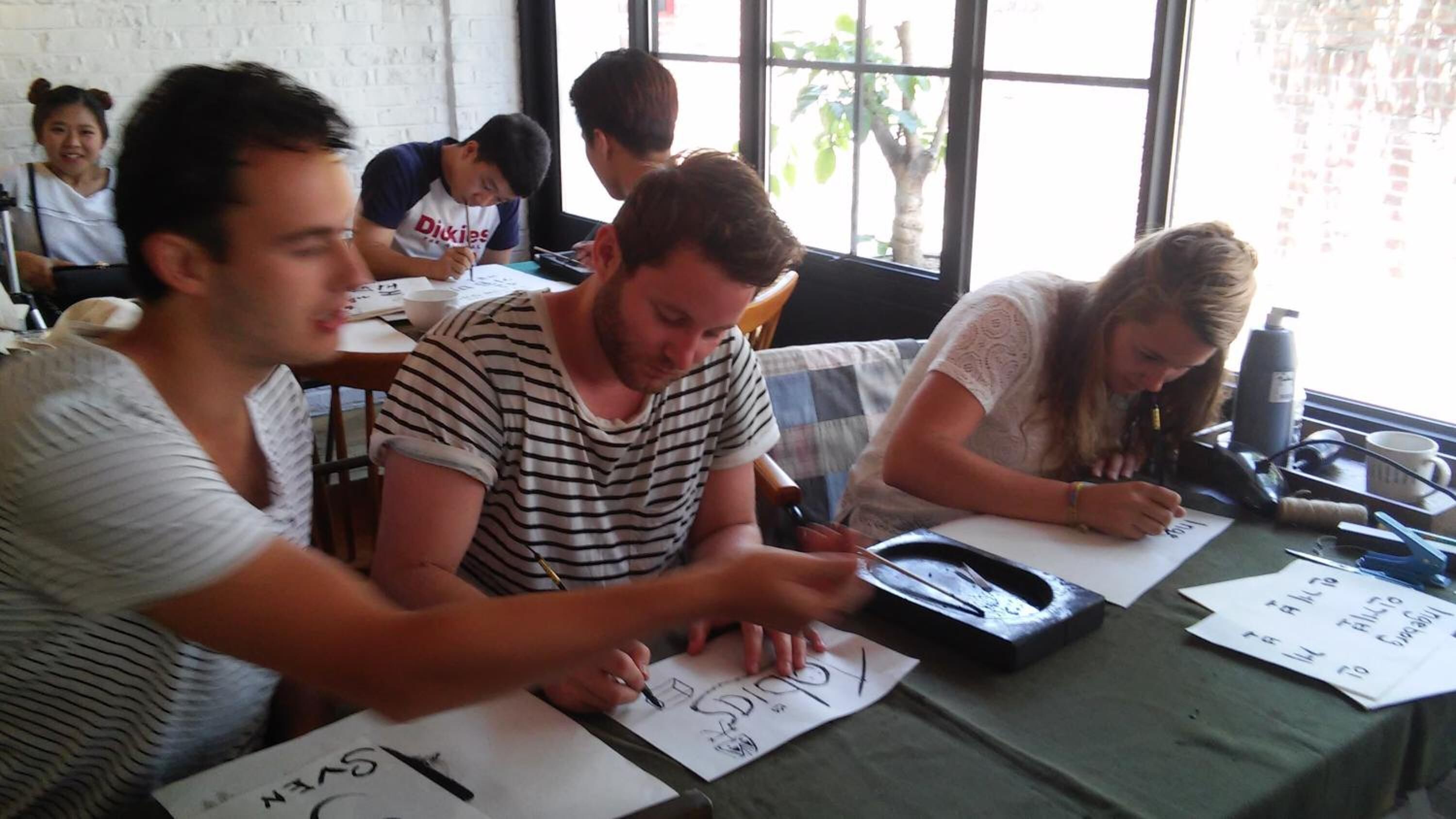 A group of people seated at tables with papers, pens, and a laptop, suggesting a meeting or workshop setting.