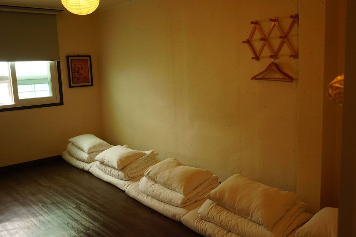 A room with a wooden floor, a window with blinds, a framed picture on the wall, and a bed with pillows.