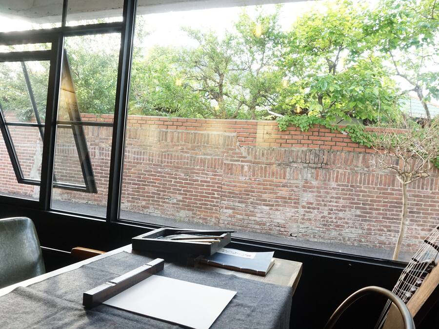 Habitación con vista a una pared de ladrillo y árboles. Se ve una ventana, una mesa con papeles y una silla.