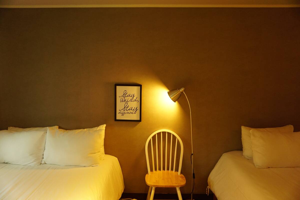A hotel room with two beds, a white chair, a wall-mounted lamp, and a framed motivational quote.