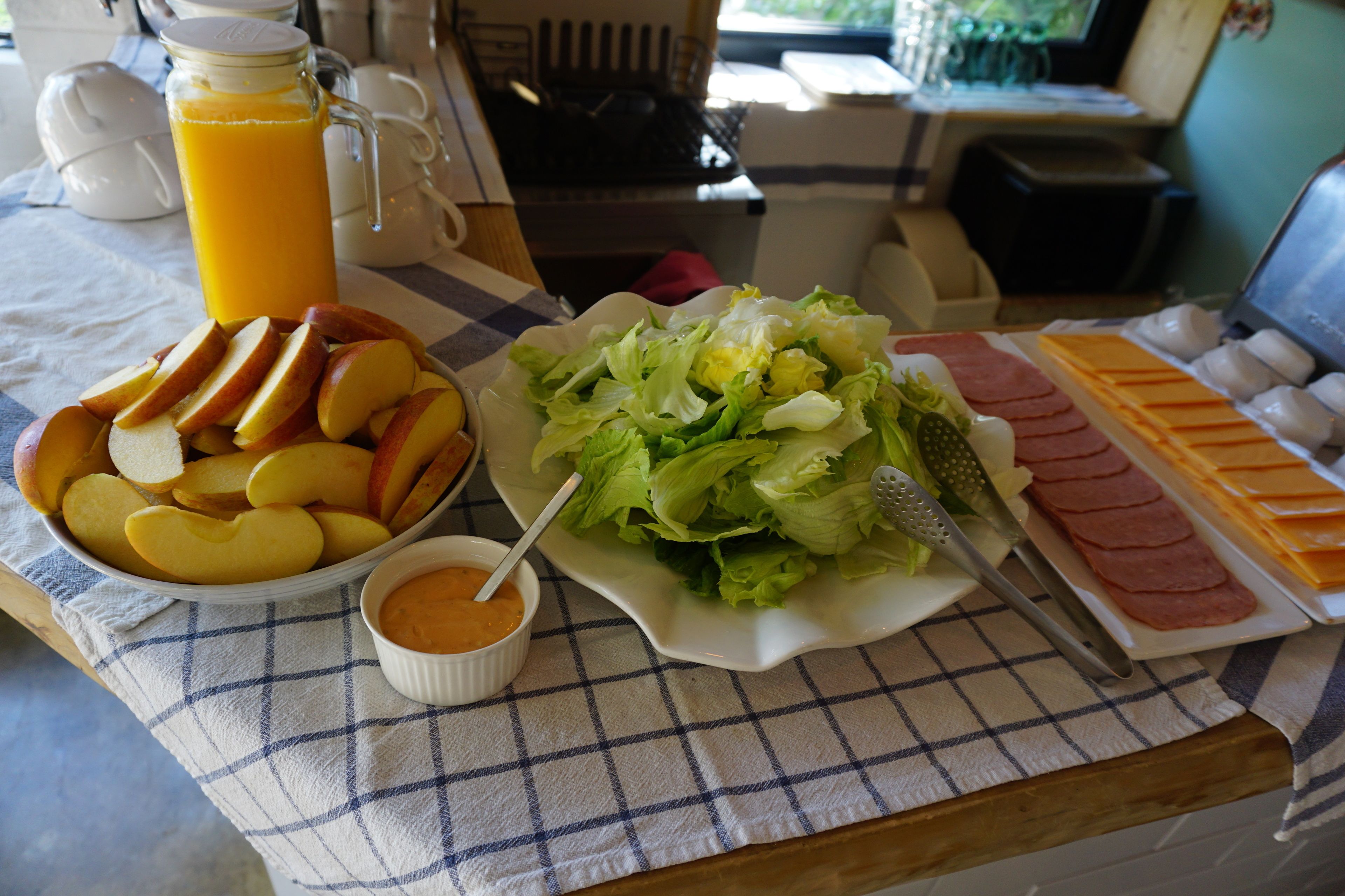A breakfast table with sliced apples, a bowl of orange juice, a salad, and sliced ham and cheese.