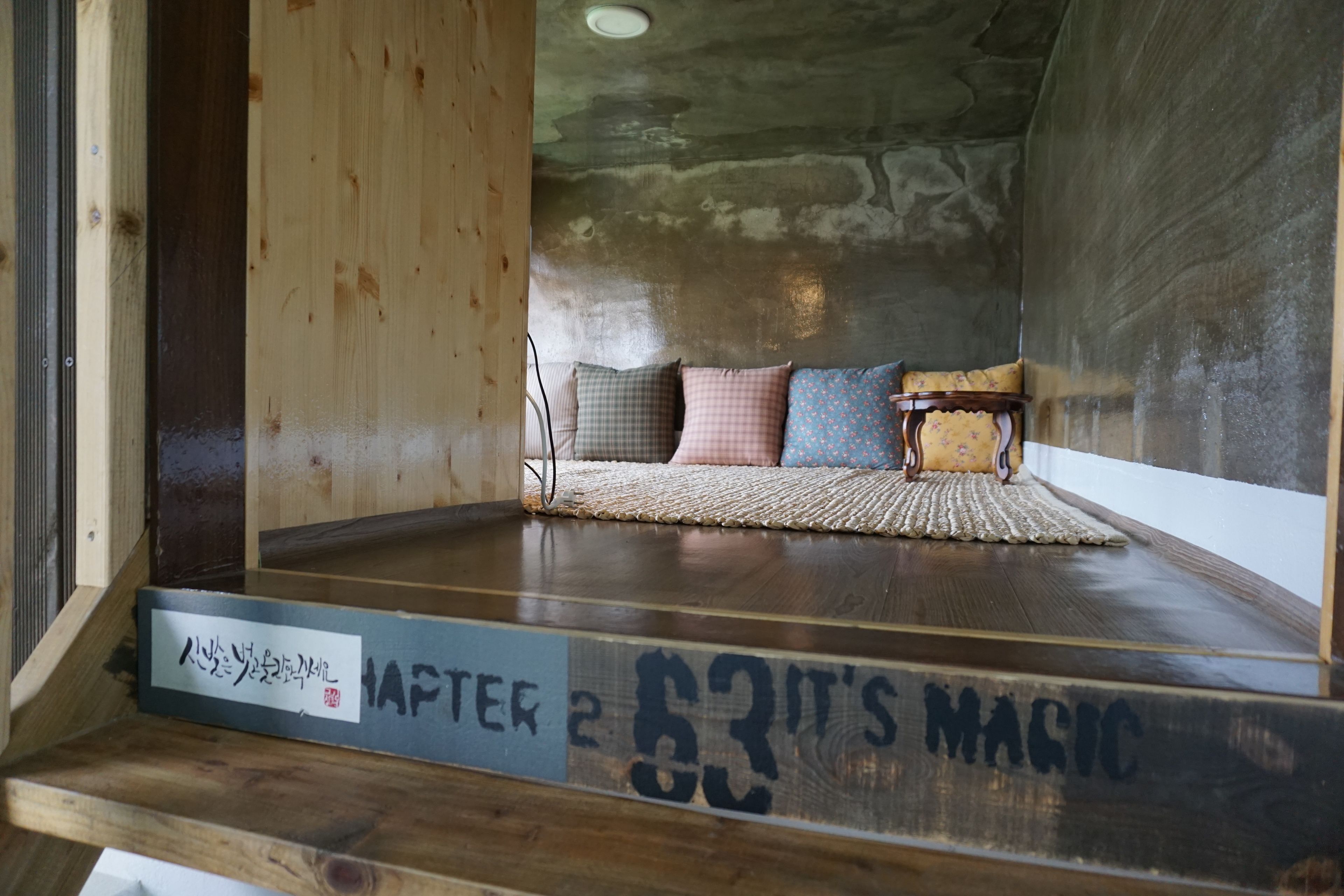 A cozy room with a wooden floor, a small sofa with cushions, and a woven rug. A book titled "AFTER 63 IT'S MAGIC" is placed on the floor.