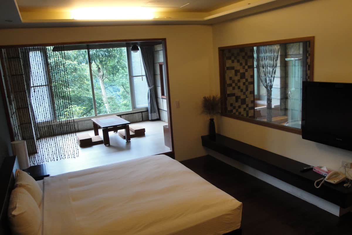 A bedroom with a bed, a TV, a table, and a window with a view of greenery.