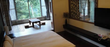 A bedroom with a bed, a TV, a table, and a window with a view of greenery.