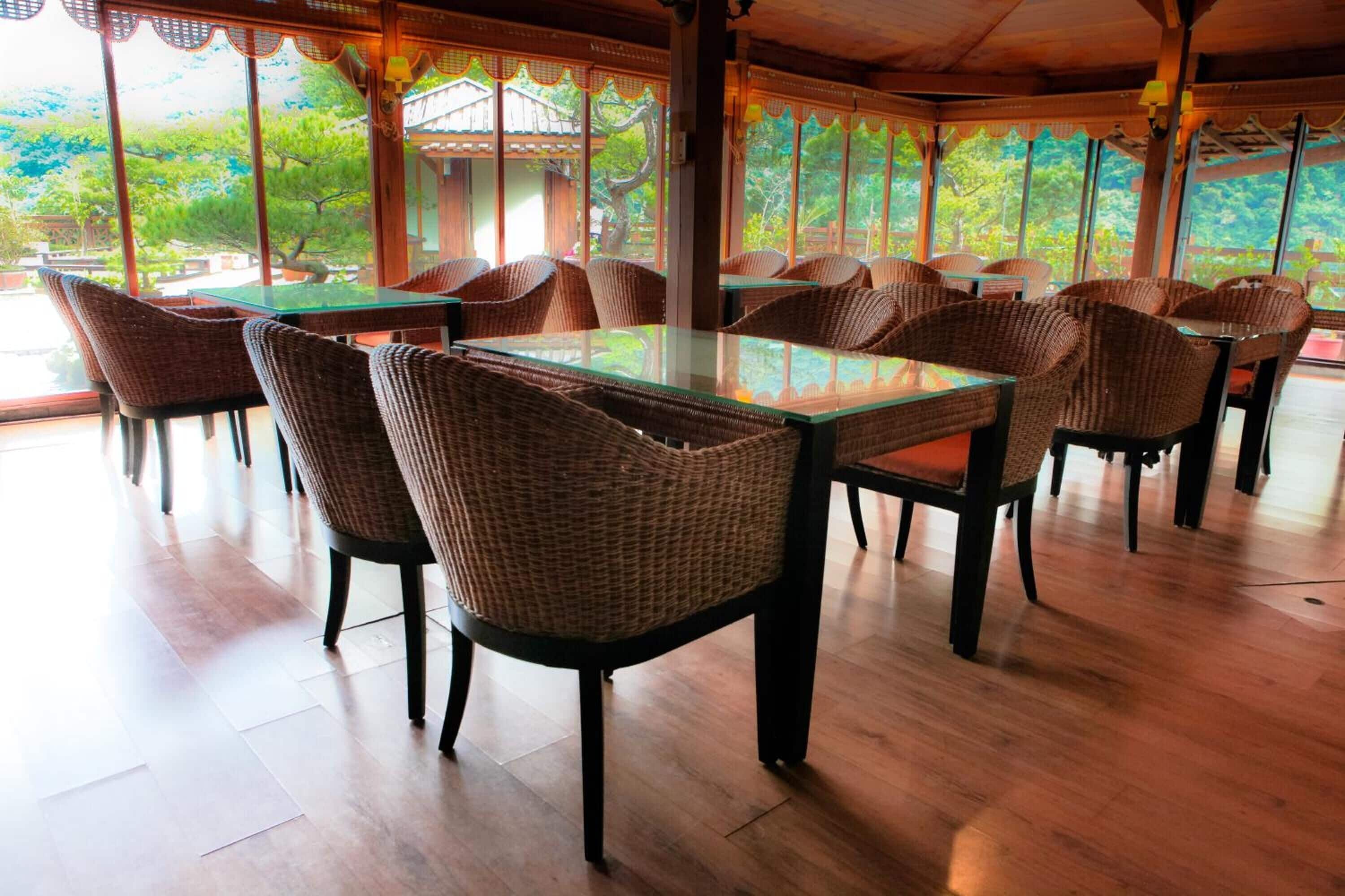 A restaurant with wicker chairs and glass tables, large windows with a view of greenery.