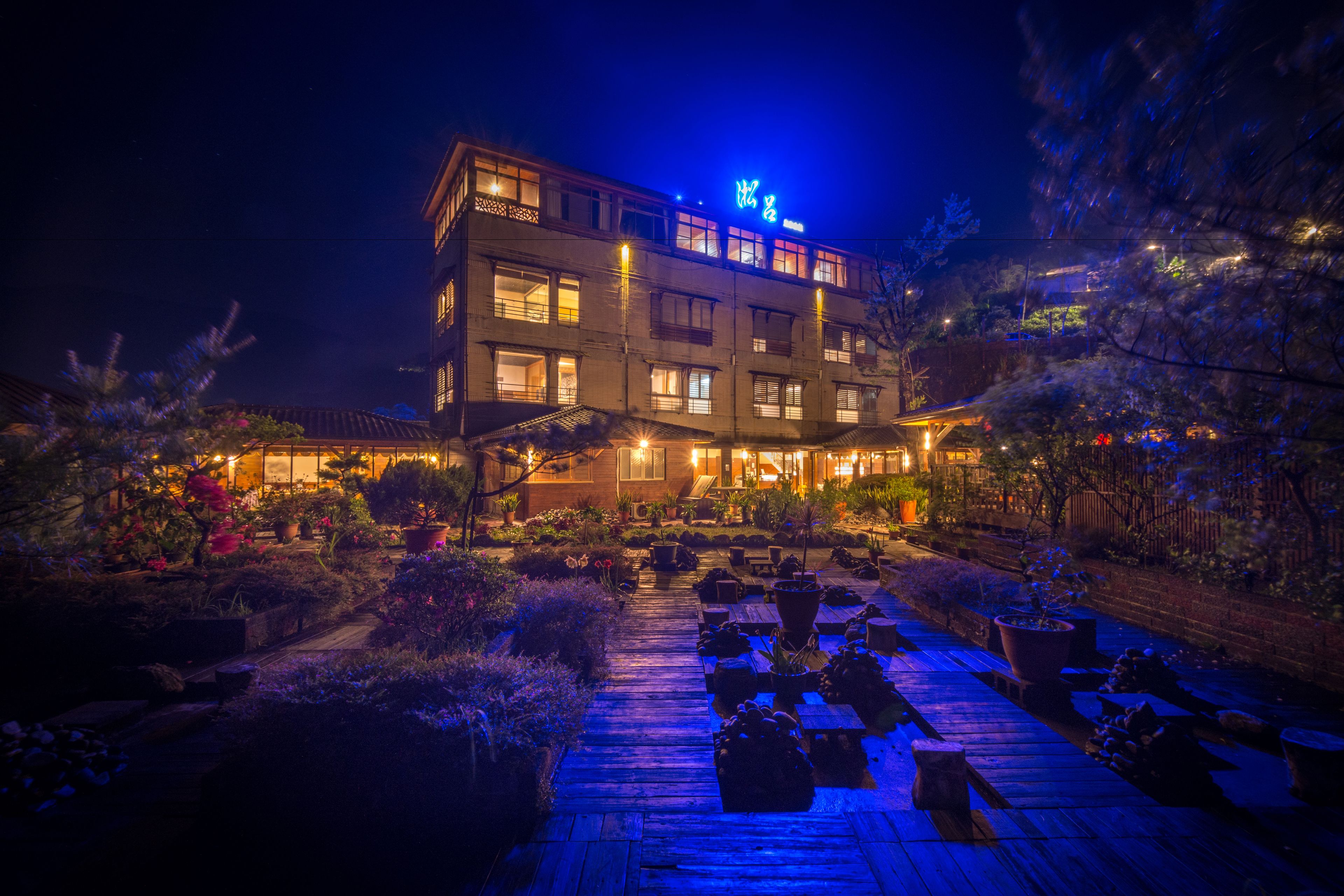 A multi-story hotel with a well-lit entrance and a garden area with potted plants and a wooden deck.