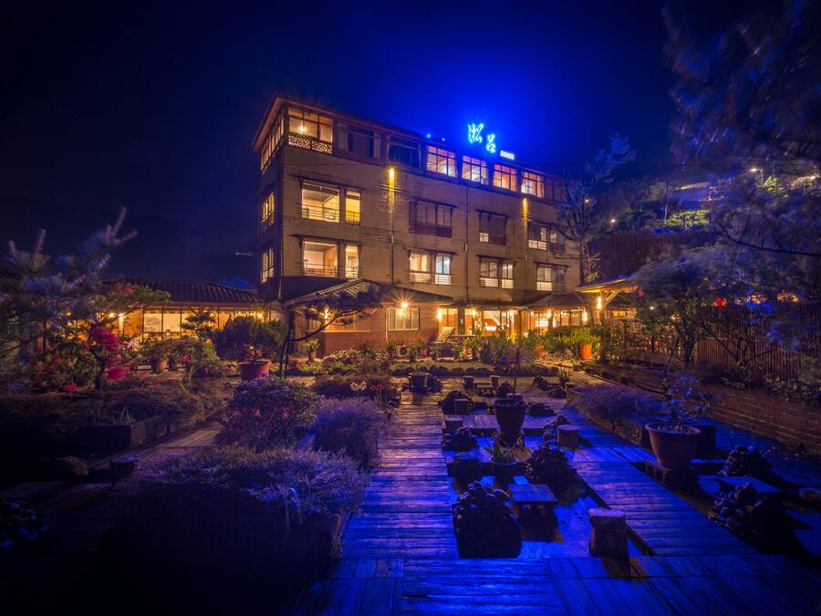 A multi-story hotel with a well-lit entrance and a garden area with potted plants and a wooden deck.