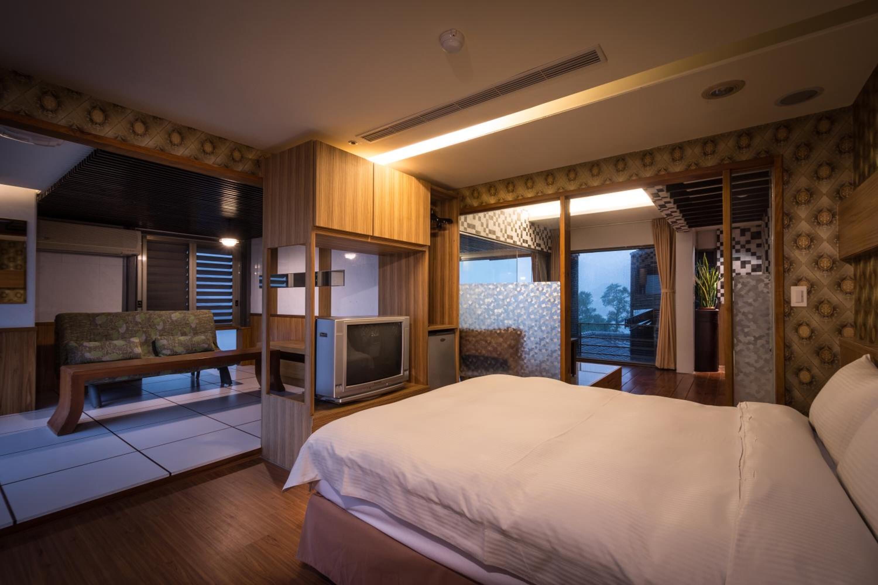A hotel room with a large bed, a television, a bench, and a view of the city through the windows.