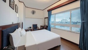 Une chambre d’hôtel avec un grand lit, un bureau, une chaise, une petite table, un téléphone, un ventilateur et une fenêtre donnant sur la ville.