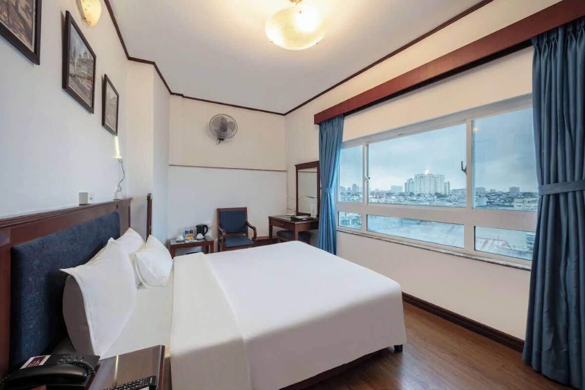 A hotel room with a large bed, a desk, a chair, a small table, a phone, a fan, and a window with a city view.