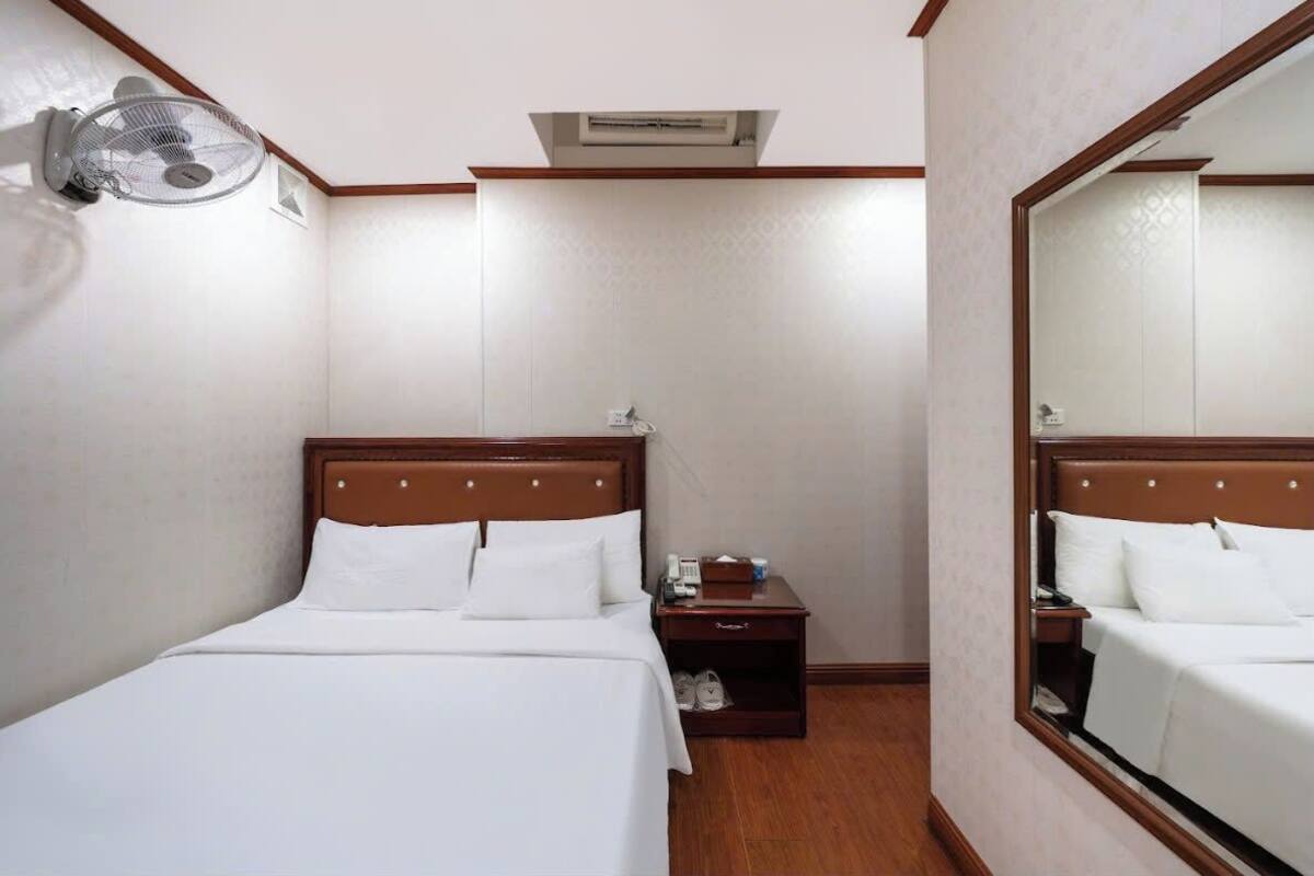 A hotel room with a bed, a nightstand, a mirror, a wall-mounted fan, and an air conditioning unit.