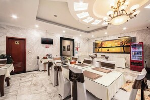 Un espace de restauration d’hôtel avec un sol en marbre, des tables blanches ornées de rubans bruns, un lustre et un tableau accroché au mur.