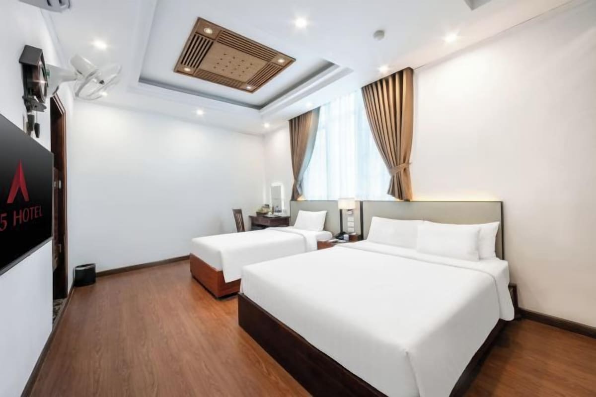Quarto de hotel com duas camas, ventilador de teto, televisão e janela com cortinas.