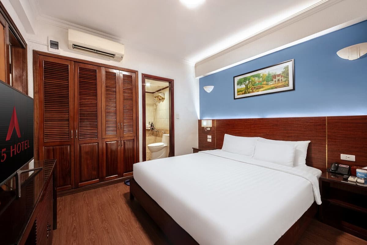 A hotel room with a large bed, wooden wardrobe, a bathroom with a toilet and shower, and a painting on the wall.