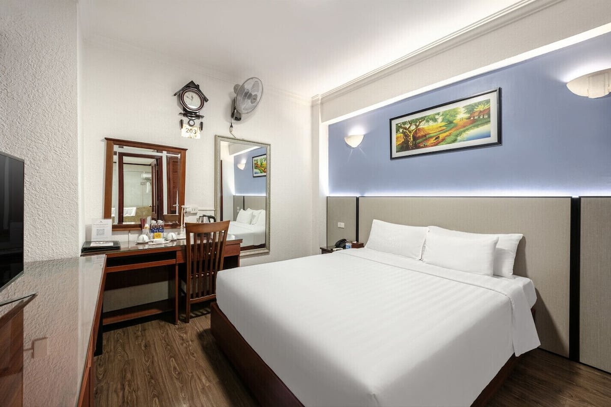 A hotel room with a large bed, a desk with a chair, a television, a clock, a mirror, and a painting on the wall.