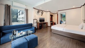 Une chambre d’hôtel avec un canapé bleu, un lit blanc, une table basse en verre, un téléviseur et une petite kitchenette.