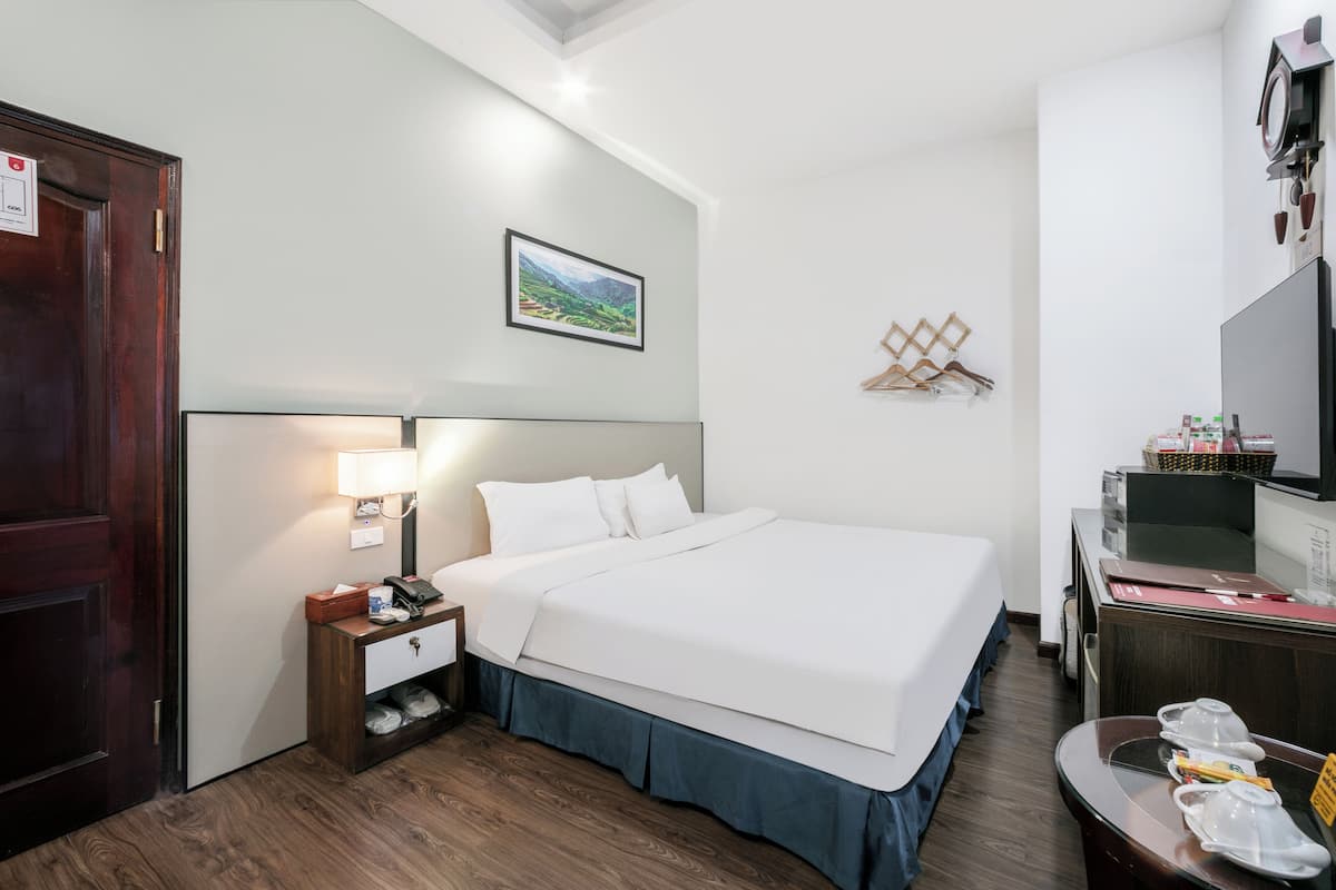 A hotel room with a bed, bedside table, lamp, and a desk with a mirror and some items on it.