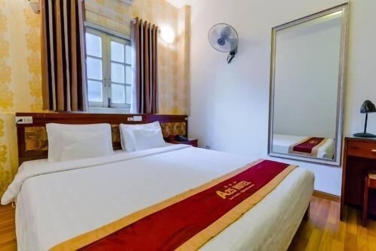 A hotel room with a bed, a window with curtains, a wall-mounted fan, and a large mirror.