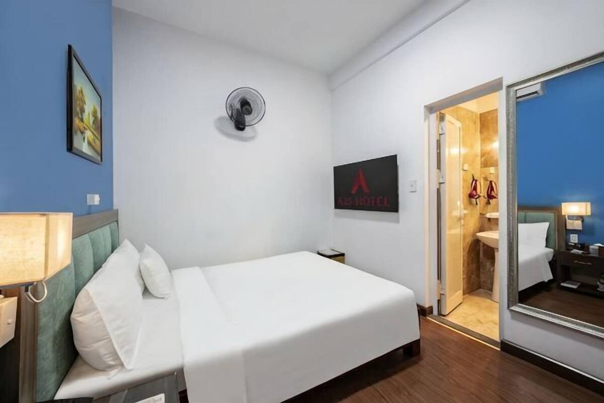 Une chambre d’hôtel comprenant un lit, une table de chevet, un miroir, un climatiseur fixé au mur et une salle de bain visible à travers une porte ouverte.
