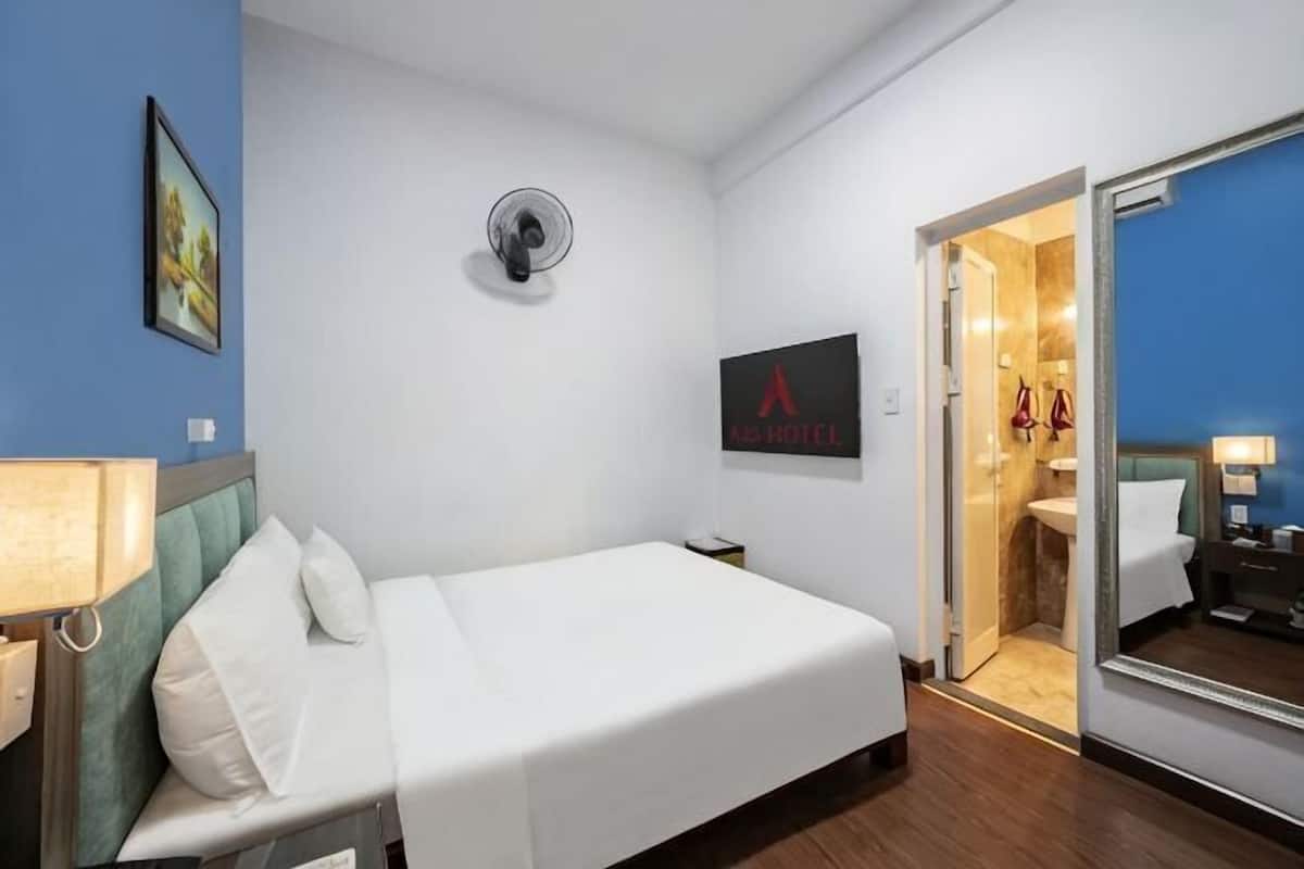 A hotel room with a bed, a bedside table, a mirror, a wall-mounted air conditioner, and a bathroom visible through an open door.