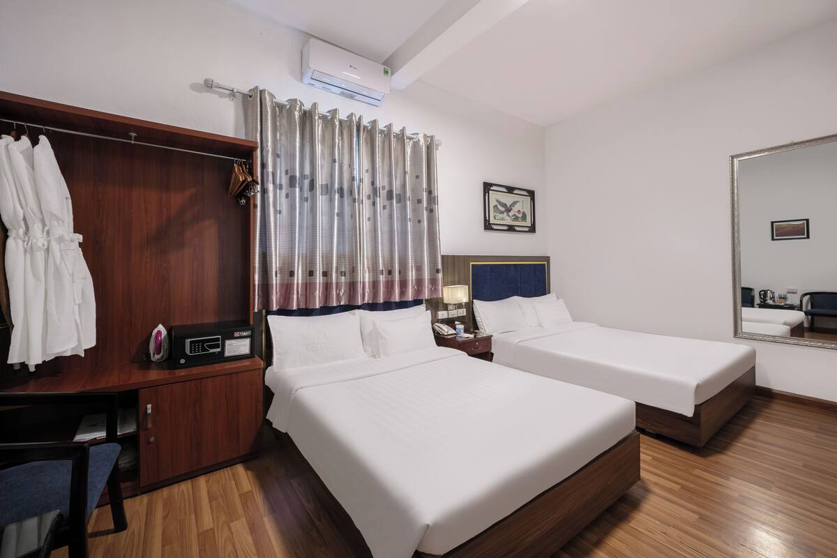 Une chambre d’hôtel avec deux lits simples, un bureau en bois avec une chaise, une armoire, un miroir et une fenêtre avec des rideaux.