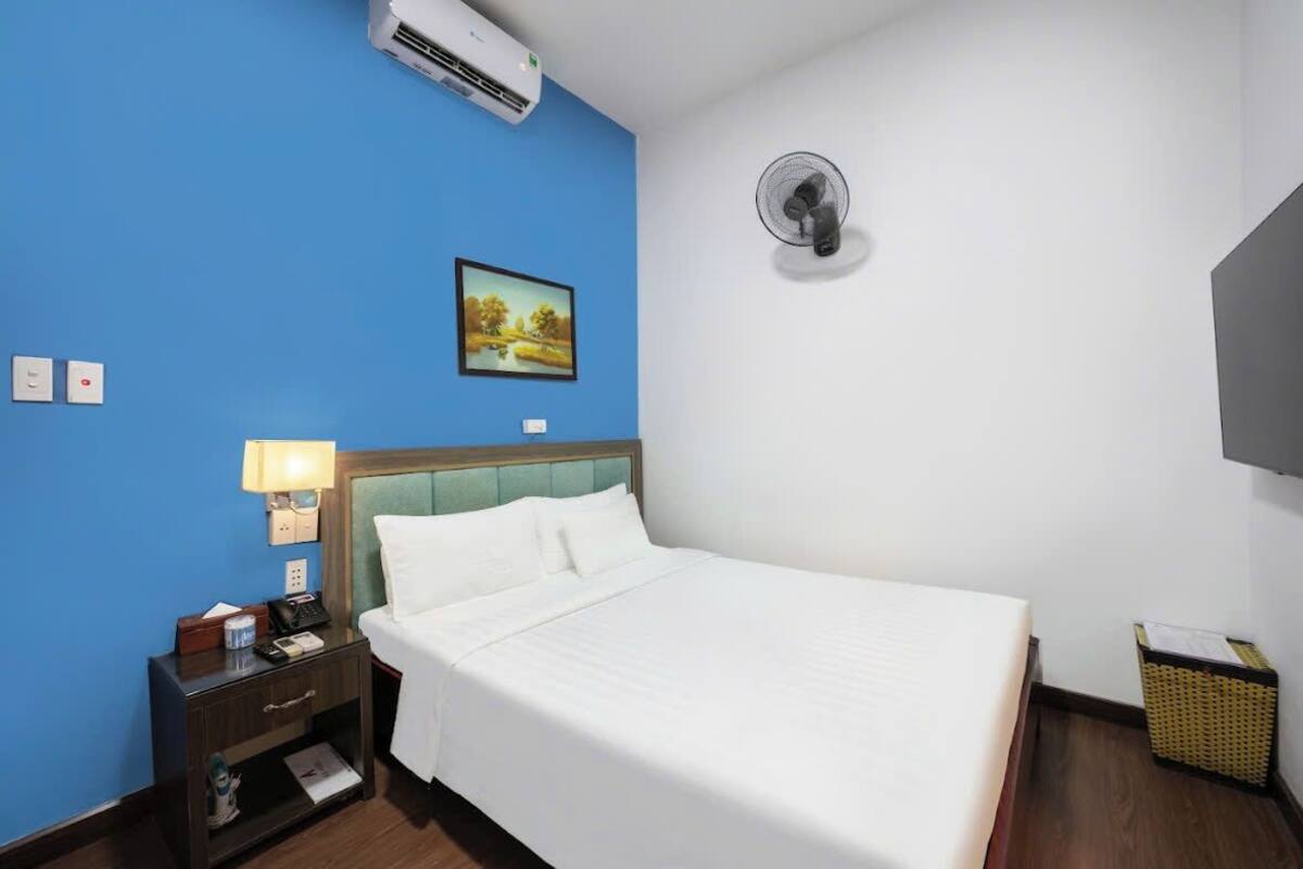 Une chambre d’hôtel comprenant un lit, une table de chevet, une lampe, un climatiseur, un ventilateur fixé au mur, un tableau et un téléviseur.