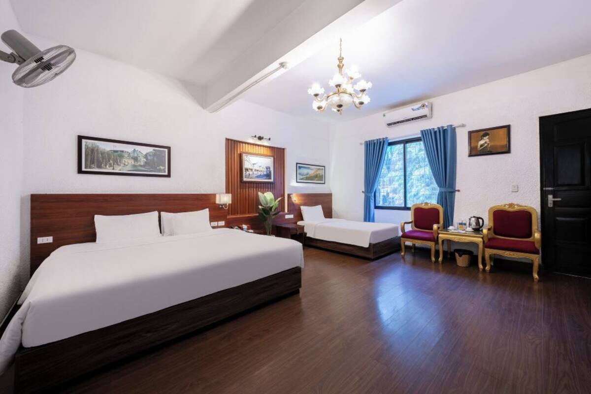 Une chambre d’hôtel avec un grand lit, deux petits lits, un bureau avec des chaises, un lustre et des tableaux encadrés aux murs.