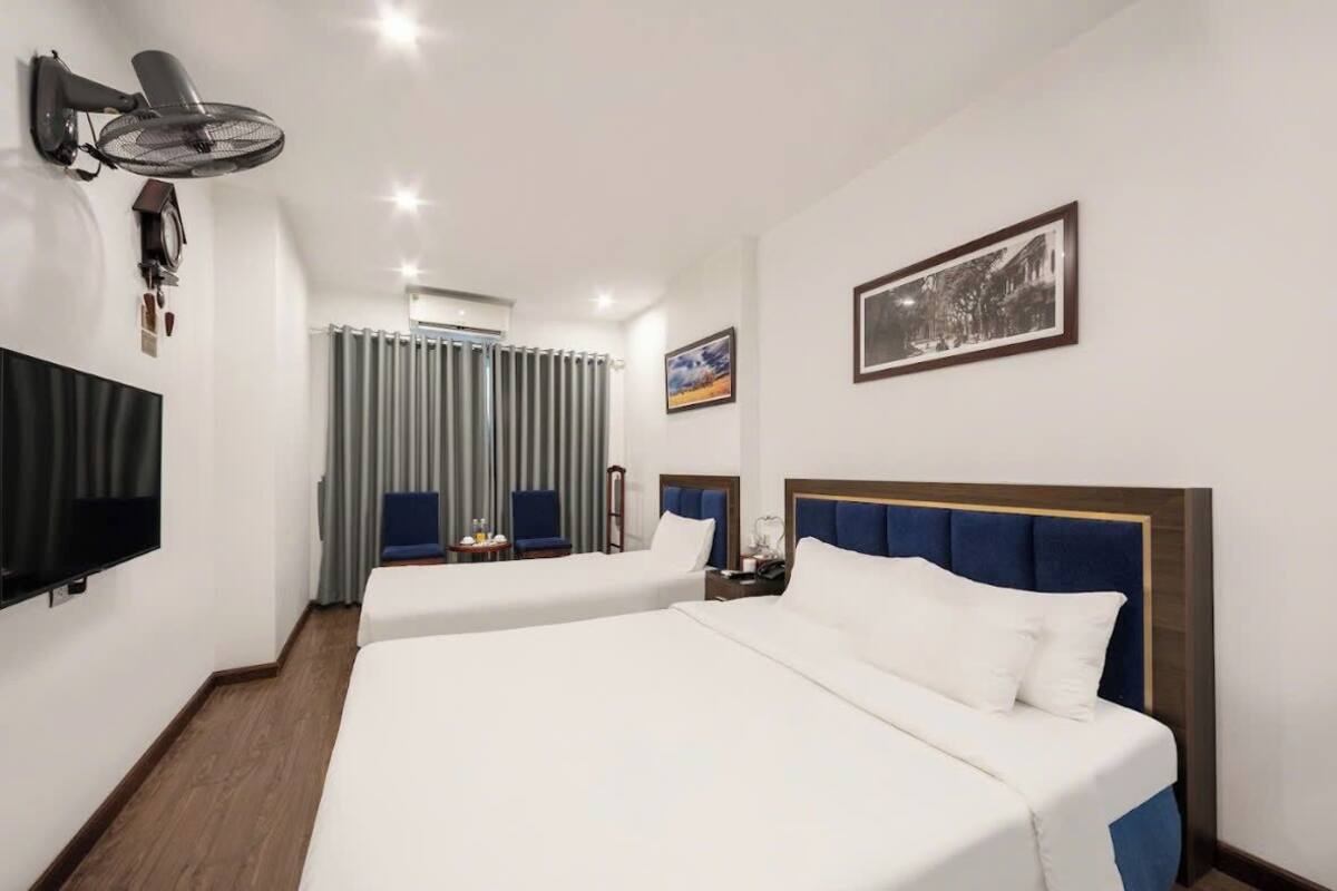 Quarto de hotel com duas camas, uma TV de tela plana, um ventilador e uma janela com cortinas, além de um quadro pendurado na parede.