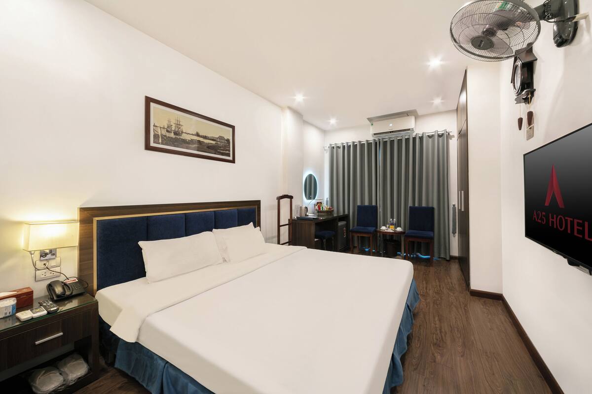 Quarto de hotel com uma cama grande, mesa de cabeceira com abajur, escrivaninha com cadeira, televisão e janela com cortinas.