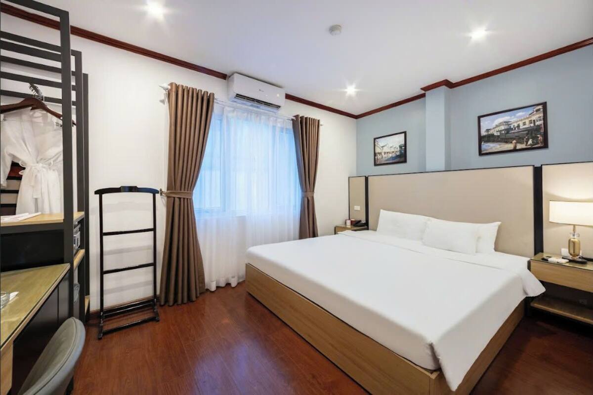 Quarto de hotel com uma cama grande, uma escrivaninha, uma cadeira, um guarda-roupa e uma janela com cortinas.