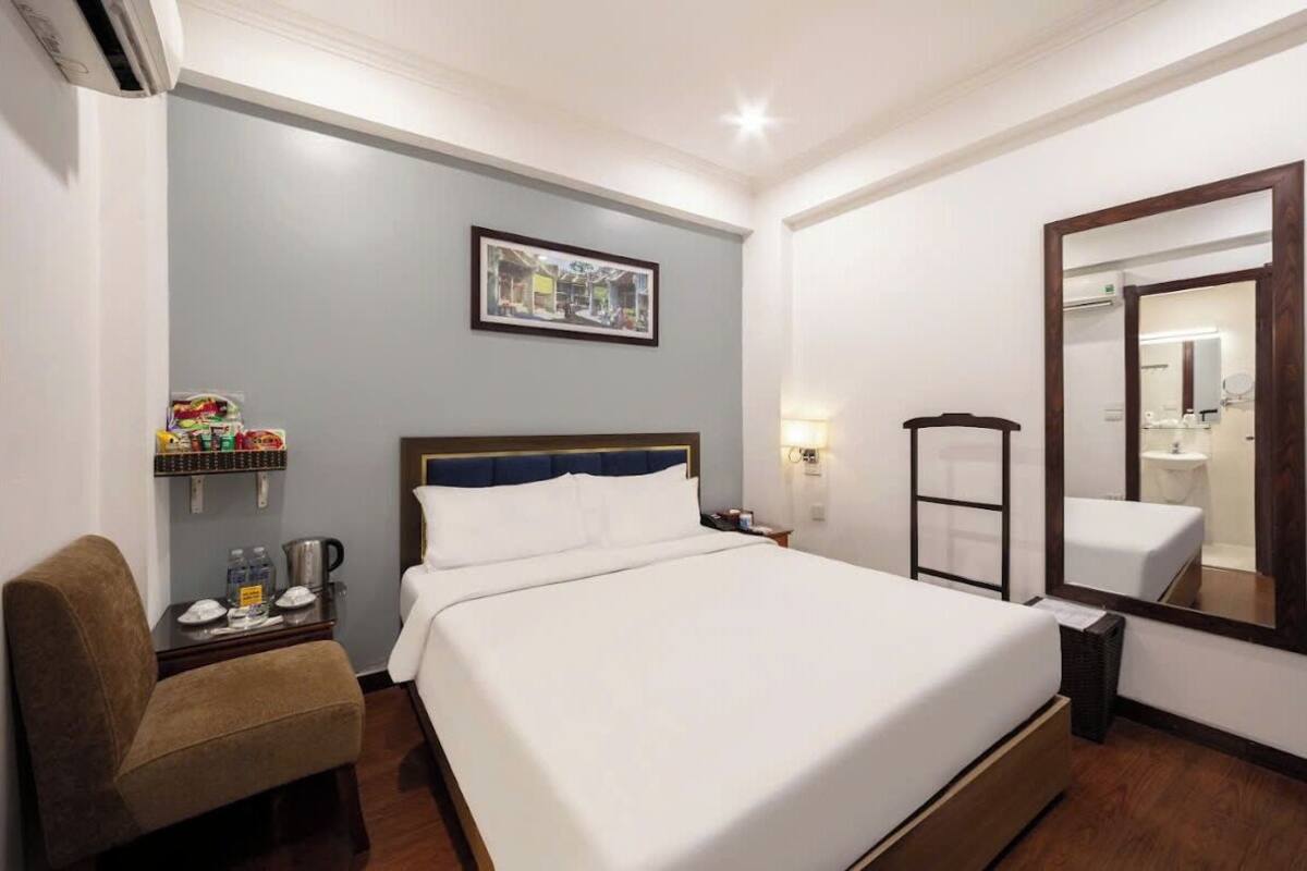 Quarto de hotel com uma cama grande, uma cadeira, uma mesinha com cafeteira, um armário com petiscos, um quadro na parede e um espelho.