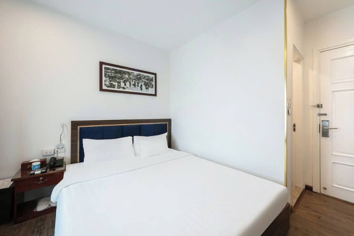 Quarto de hotel com uma cama grande, uma mesa de cabeceira com abajur, um quadro na parede e uma porta à direita.