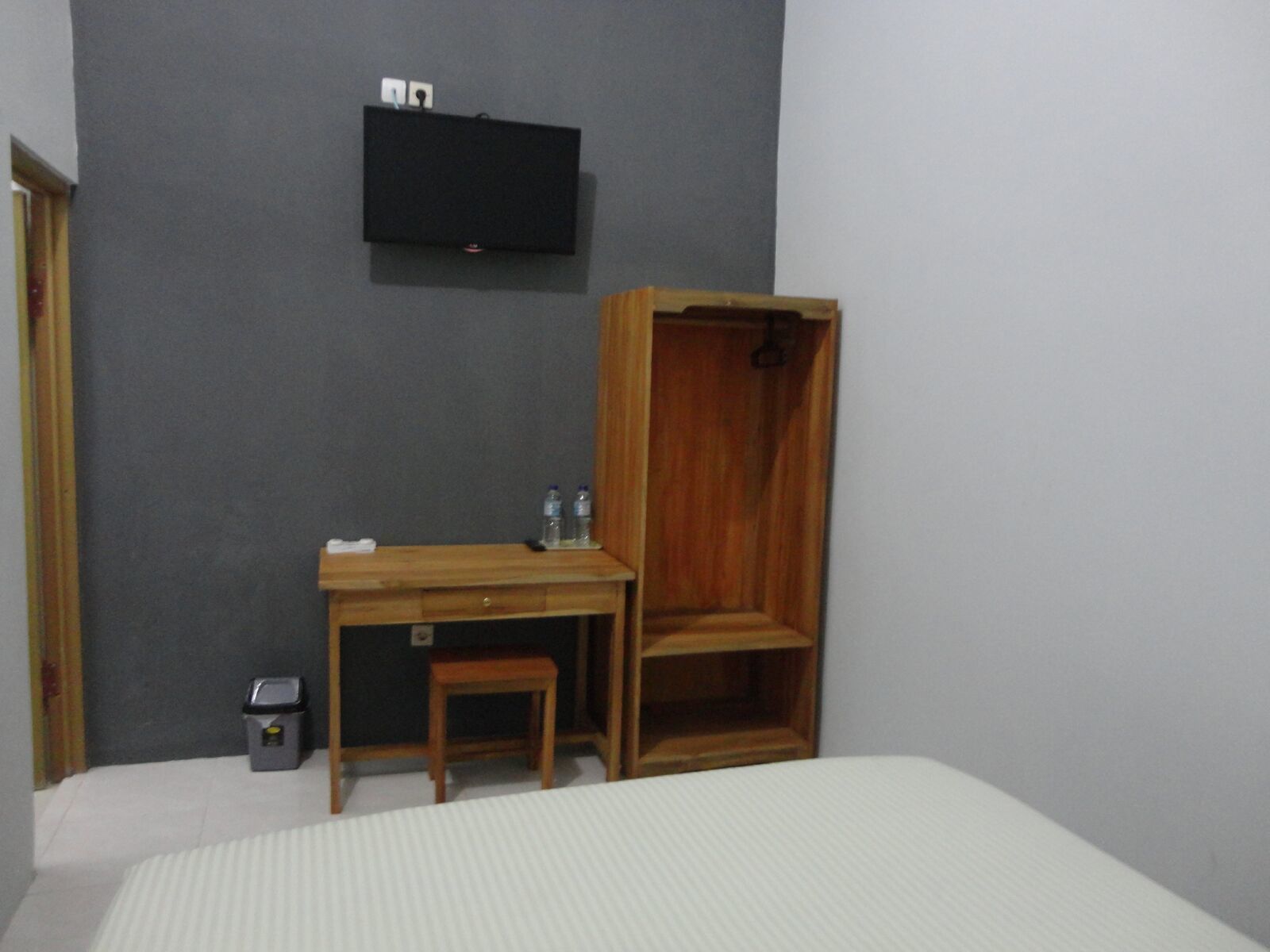 A bedroom with a bed, a desk with a chair, a wardrobe, a small trash bin, and a mounted TV.