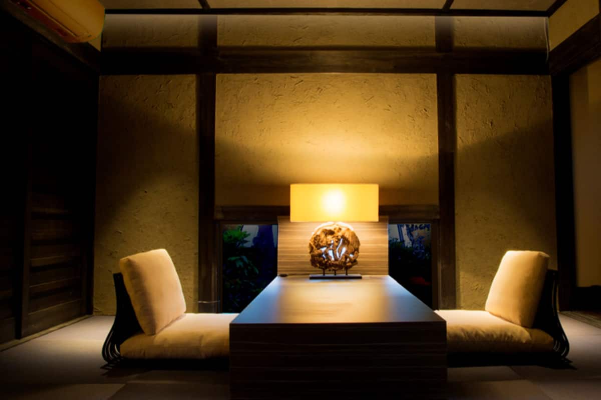 Ein Zimmer mit einem zentralen Tisch, zwei Stühlen, einer Lampe und einem Wandregal mit einem dekorativen Gegenstand.