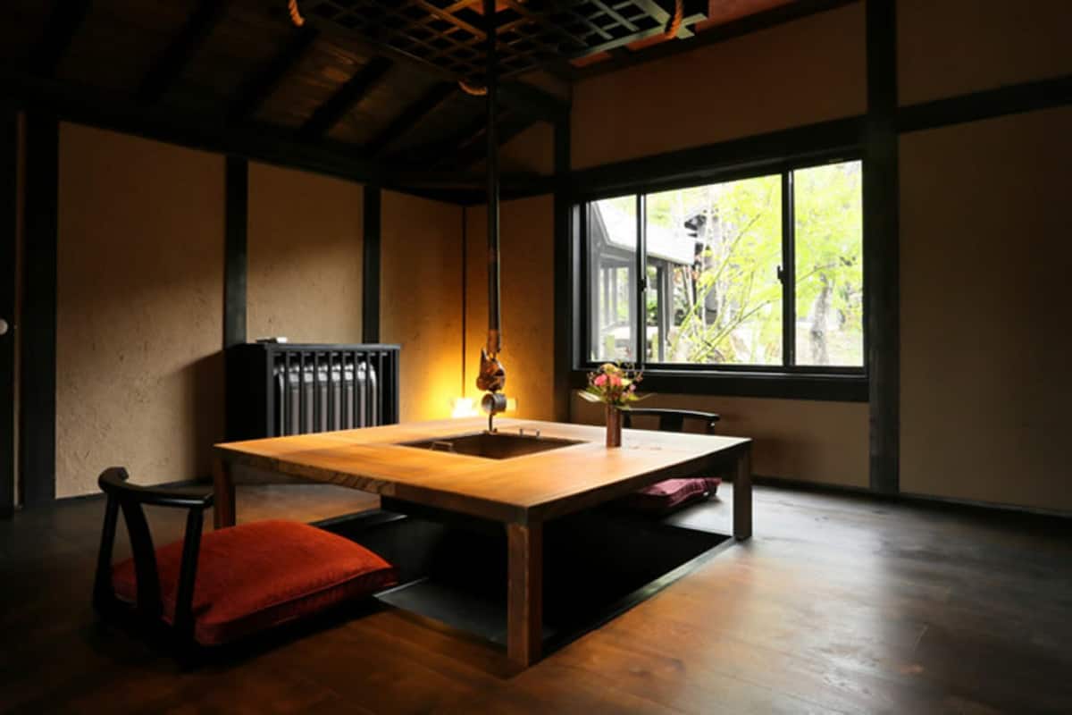 Ein traditioneller japanischer Raum mit Tatami-Böden, einem niedrigen Holztisch, einem roten Kissen und einem Fenster mit Blick auf Grünflächen.