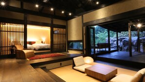 Una habitación tradicional japonesa con suelo de tatami, una cama baja de madera, una zona de estar con cojines y vistas a un jardín rocoso a través de amplios ventanales.