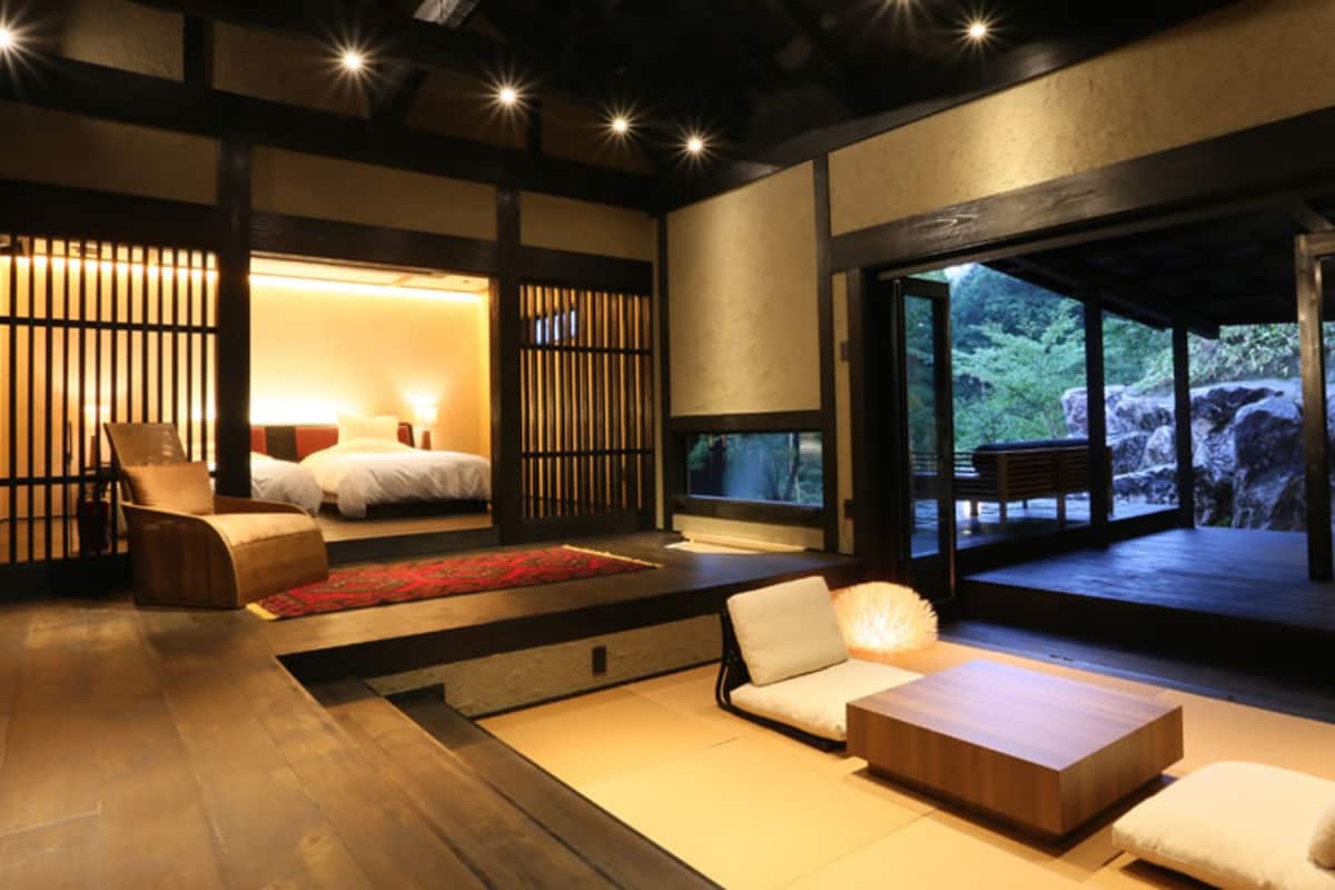 Ein traditioneller japanischer Raum mit Tatami-Böden, einem niedrigen hölzernen Bett, einer Sitzecke mit Kissen und Blick auf einen Felsengarten durch große Fenster.