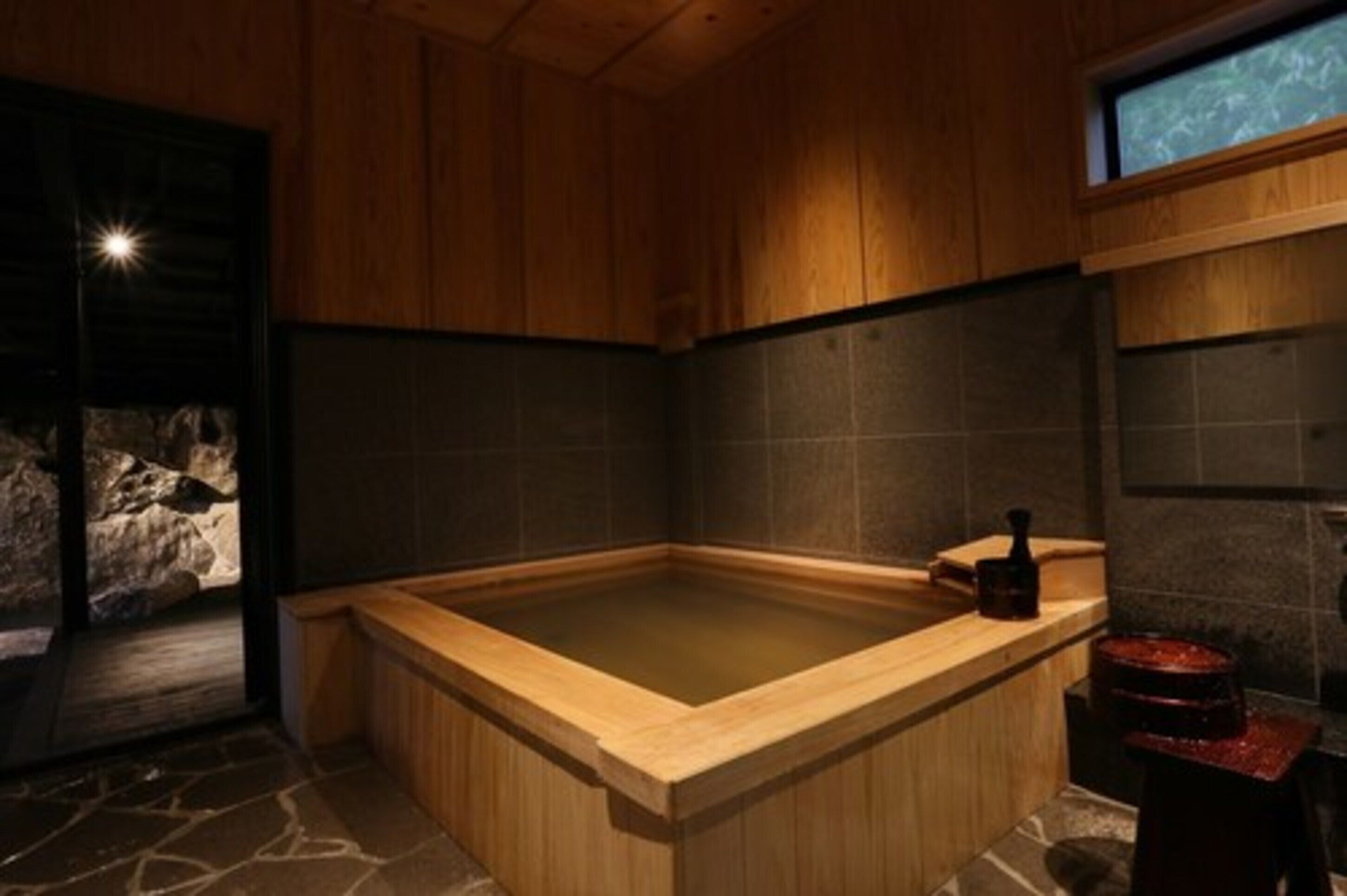 A wooden-floored hot tub with a stone wall backdrop and a small stool.