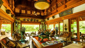 A spacious room with wooden beams, a large window, and a variety of furniture including a sofa, chairs, and a table with books. - Howie's Homestay (Mae Rim)