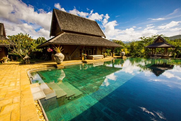 A pool area with a clear blue pool, a wooden structure with a sloped roof, and surrounding greenery.