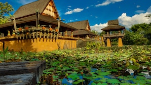 A traditional wooden building with a thatched roof, surrounded by a pond with lily pads. - Howie's Homestay (Mae Rim)