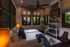 A bedroom with a large bed, a ceiling fan, a nightstand with a lamp, a shelf with decorative items, and a window with a view of greenery. - Howie's Homestay (Mae Rim)
