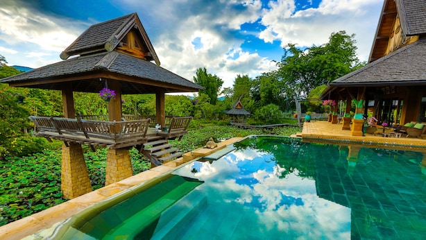 A clear swimming pool with wooden deck chairs and a gazebo, surrounded by lush greenery. - Howie's Homestay (Mae Rim)